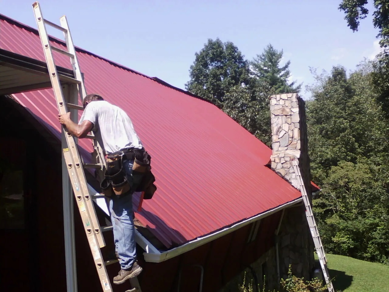 Roofer on a ladder beside a red metal roof and stone chimney — Soffit & Fascia Repair by Roofing Brothers in Malden On Hudson